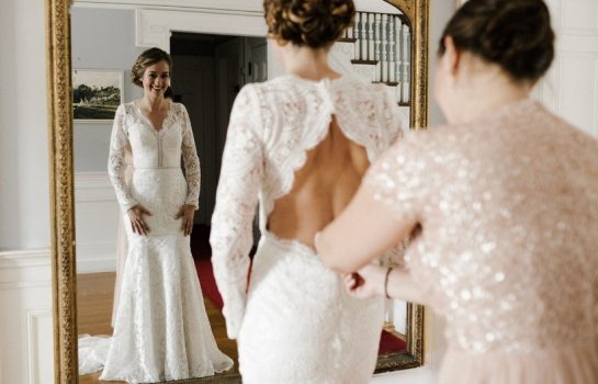 a girl wearing a wedding and standing in front of mirror another girl helping